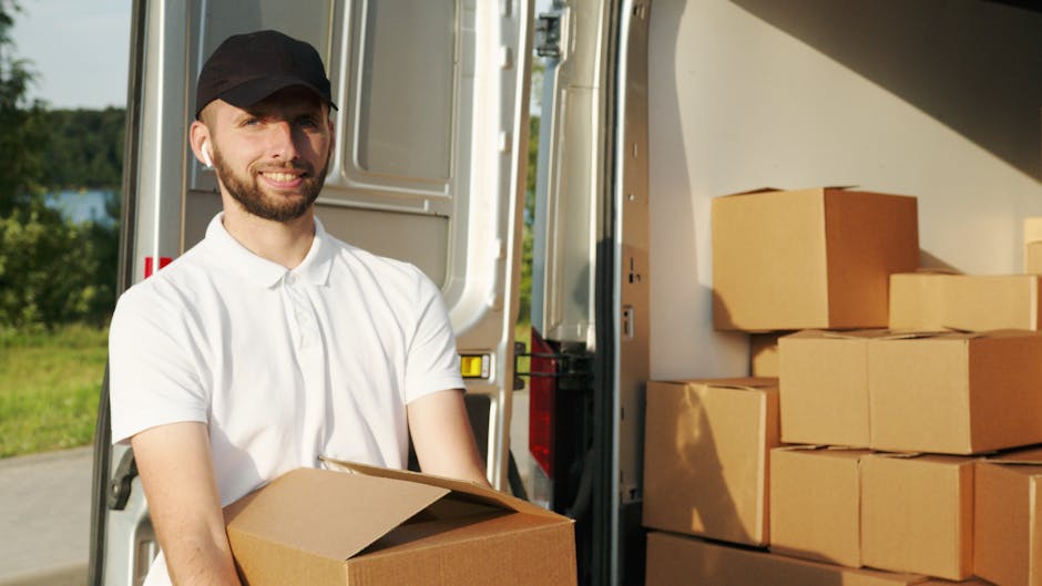 A man with a beard, wearing a white polo shirt and a black cap, standing outside near the open back of a white moving van, is holding a cardboard box for home relocation. The van is filled with severa