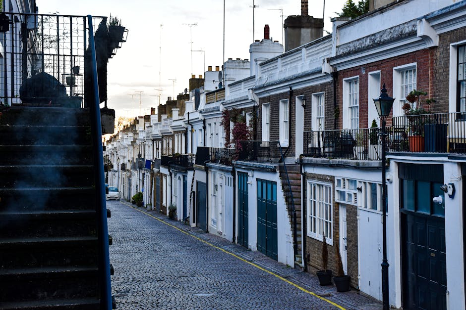 The image depicts a narrow residential street lined with terraced houses featuring brick and white-painted facades, small front gardens with potted plants, and black wrought-iron balconies. A set of e